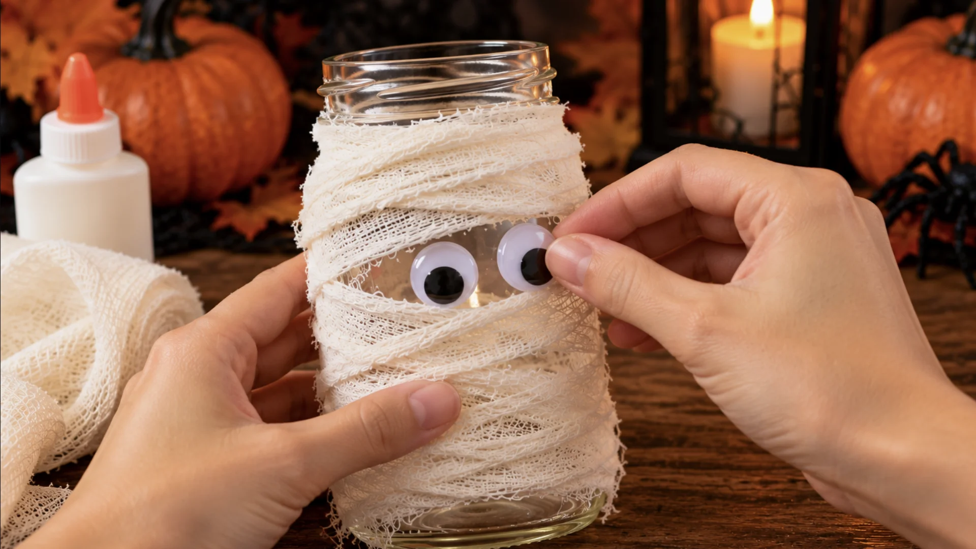 Mãos colando olhos móveis em pote de vidro envolvido com gaze para criar uma múmia de Halloween.