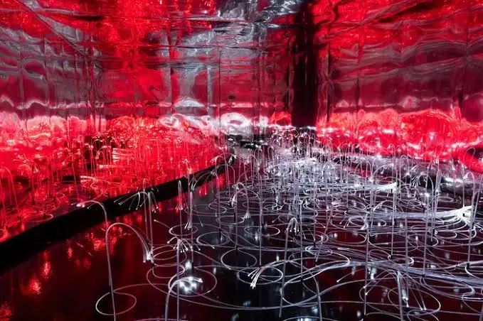 Instalação artística com luzes vermelhas, espelhos e estruturas luminosas.
