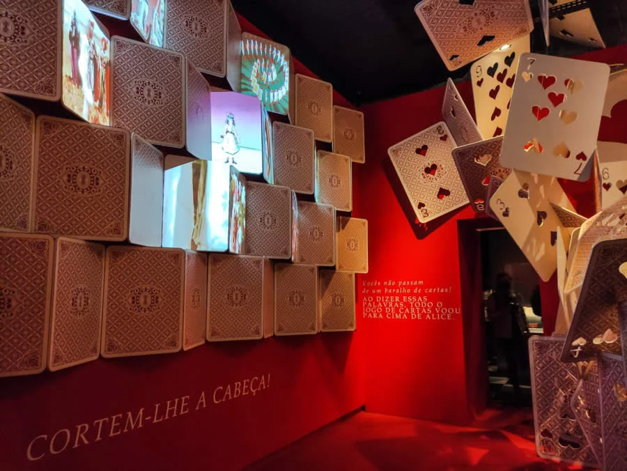 Instalação imersiva de Alice no País das Maravilhas com cartas gigantes e projeções.