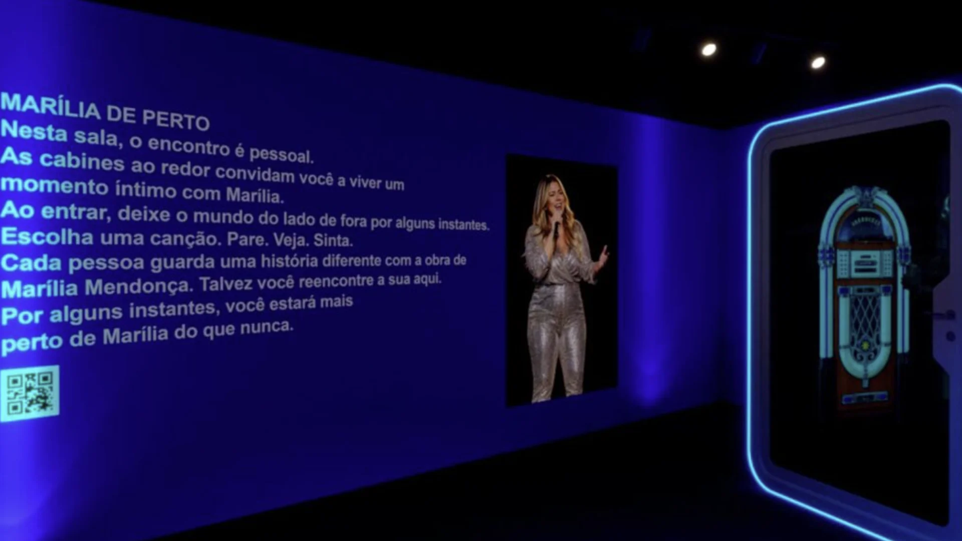 Sala interativa da exposição Marília Mendonça no MIS, com iluminação azul, imagem da cantora e cabine musical.