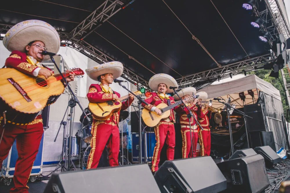Grupo de mariachis com trajes vermelhos e sombreros mexicanos se apresenta em palco com instrumentos musicais.