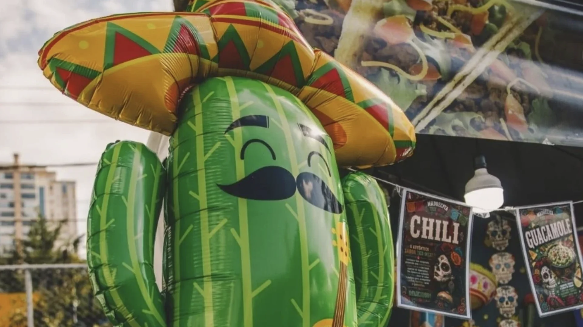 Cacto inflável verde com sombrero mexicano diante de barraca com cartazes de comidas típicas e caveiras decorativas.