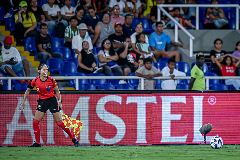 Árbitra assistente em campo durante partida de futebol feminino, com publicidade da Amstel ao fundo.