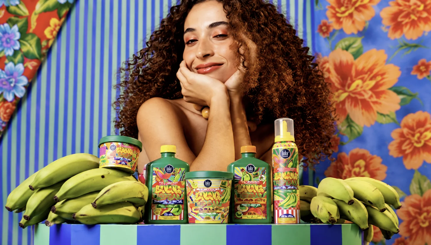 Modelo de cabelos cacheados posa com produtos Banana Tropicana da Lola Cosmetics em cenário colorido.