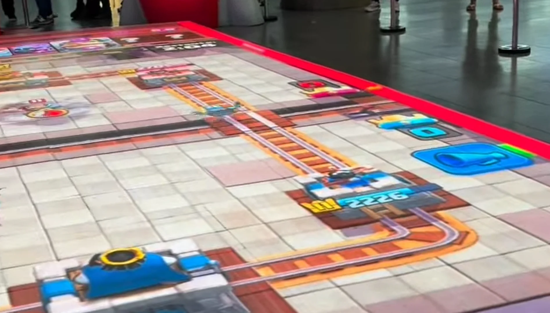 Arena de Clash Royale reproduzida em um grande painel no piso, com torres, trilhos e elementos do jogo.